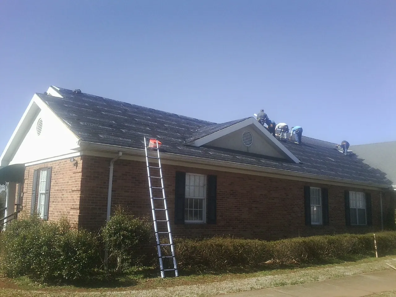 Crew tearing shingles off a brick ranch roof with a ladder set — Emergency Roof Repair work by our crew in Milan